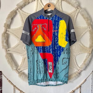 Könnte beinhalten: Ein kurzärmeliges Radtrikot mit einem lebendigen, abstrakten Design. Das Trikot hat einen grauen Kragen und Ärmel, einen blauen Körper mit roten, gelben und orangen Formen und einen teilweisen Reißverschluss. Das Wort "ACTIVE" ist auf dem Ärmel zu sehen.