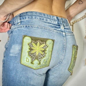 May include: Light blue denim jeans with decorative, embroidered pockets. The pockets feature a green and brown floral design with intricate detailing. The jeans have a classic five-pocket design and a button and zip closure.