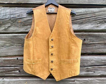 May include: A tan suede vest with a V-neck, button closure, and two front pockets. The vest is hanging on a black hanger against a rustic wooden background. The vest has a small decorative label near the collar.