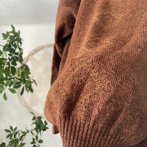 May include: Close-up of a rust-colored sweater with a ribbed hem and cuffs. The knitwear has a textured appearance with subtle variations in color, creating a warm and cozy aesthetic. The sweater's design suggests a relaxed fit.