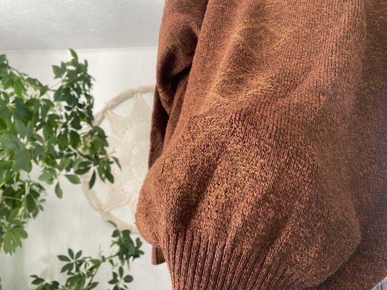 May include: Close-up of a rust-colored sweater with a ribbed hem and cuffs. The knitwear has a textured appearance with subtle variations in color, creating a warm and cozy aesthetic. The sweater's design suggests a relaxed fit.