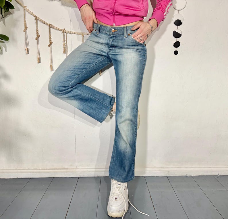 May include: Faded blue denim jeans with a slight flare at the ankle. The jeans have a classic five-pocket design and a button and zipper closure. The jeans are worn with white sneakers.