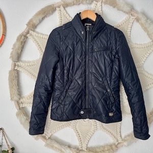 May include: A navy blue quilted jacket featuring a high collar, a full-length front zipper, and a small chest pocket. The jacket has long sleeves, a snap button closure at the hem, and a subtle textured diamond pattern across the entire surface.