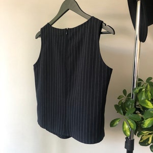 Puede incluir: Una blusa sin mangas de rayas blancas y negras con cierre de cremallera en la espalda. La blusa est&aacute; colgada de una percha negra.