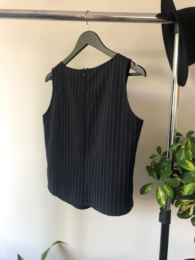 Puede incluir: Una blusa sin mangas de rayas blancas y negras con cierre de cremallera en la espalda. La blusa est&aacute; colgada de una percha negra.