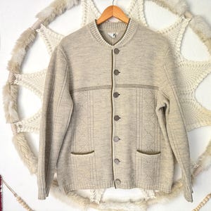 May include: A beige knit cardigan featuring a round neckline, long sleeves, and a front button closure. The garment includes two front pockets and a subtle diamond-patterned texture on the lower section, finished with a dark trim along the placket.