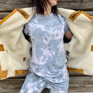 May include: A brown suede jacket with a white sherpa lining. The jacket has a zipper closure and a button-up front. The jacket is being held open by a person wearing a blue tie-dye shirt.