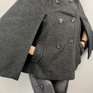 May include: A charcoal gray cape coat with a double-breasted design. The coat features large, round, brown buttons and two front pockets. The sleeves are designed as capes, with openings for the arms.