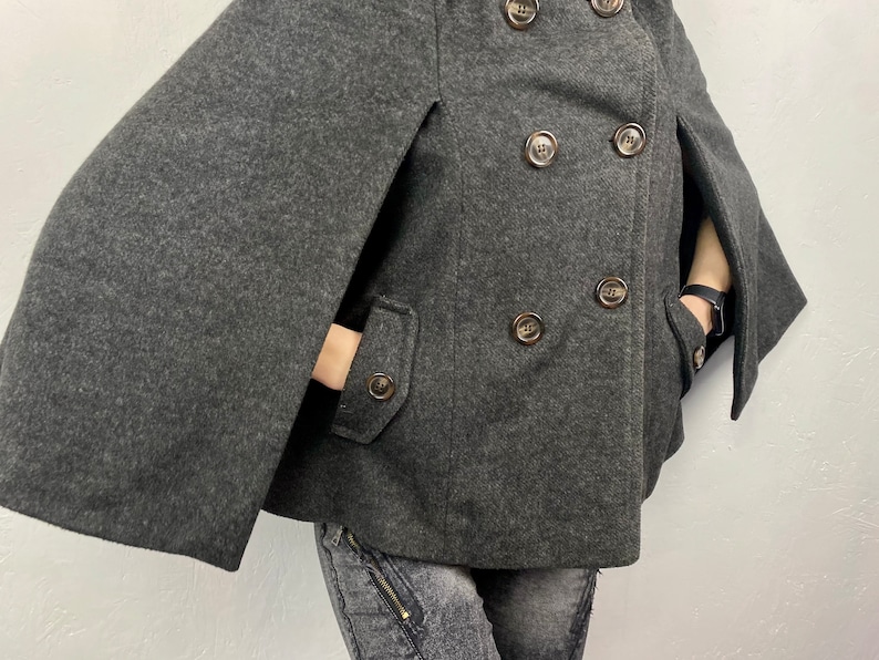 May include: A charcoal gray cape coat with a double-breasted design. The coat features large, round, brown buttons and two front pockets. The sleeves are designed as capes, with openings for the arms.