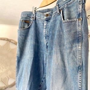 May include: Light blue denim jeans hanging on a wooden hanger. The jeans have classic five-pocket styling, a button and zipper closure, and a straight leg fit. The denim has a faded wash, giving it a vintage look.