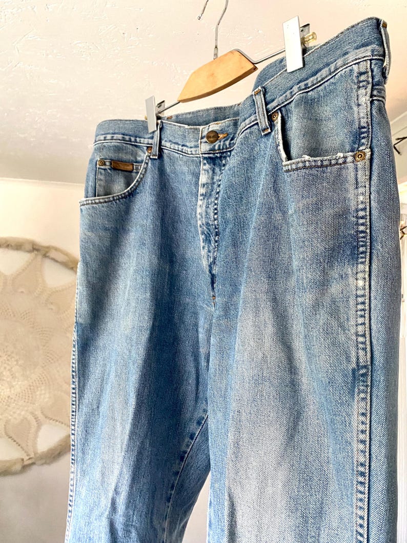 May include: Light blue denim jeans hanging on a wooden hanger. The jeans have classic five-pocket styling, a button and zipper closure, and a straight leg fit. The denim has a faded wash, giving it a vintage look.