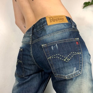 May include: A pair of blue denim jeans with a brown leather patch that says "Famous Quality Replay" on the back. The jeans have a light green stitching detail on the back pockets.