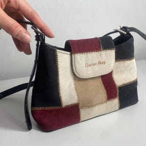 May include: A small, patchwork shoulder bag with a black strap. The bag features a flap with the text "Daniel-Ray" and is made of suede in black, cream, tan, and burgundy colours.