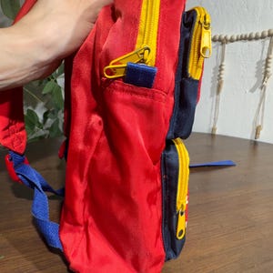 May include: A red backpack with yellow zippers and blue accents. The backpack has two zippered compartments and adjustable blue shoulder straps. The bag is made of a shiny, durable material.