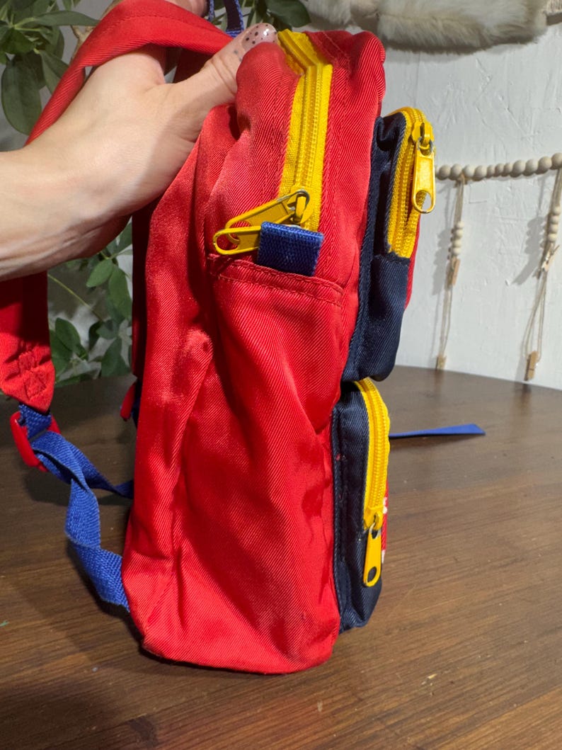 May include: A red backpack with yellow zippers and blue accents. The backpack has two zippered compartments and adjustable blue shoulder straps. The bag is made of a shiny, durable material.