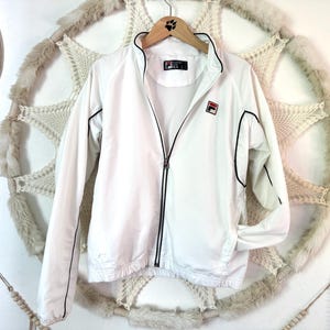 May include: White Fila track jacket with black trim and a full-length zipper. The jacket features the Fila logo on the chest and a small logo on the collar. The jacket is hanging on a wooden hanger.