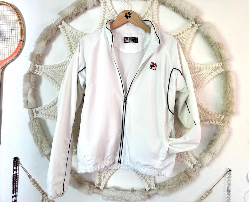 May include: White Fila track jacket with black trim and a full-length zipper. The jacket features the Fila logo on the chest and a small logo on the collar. The jacket is hanging on a wooden hanger.