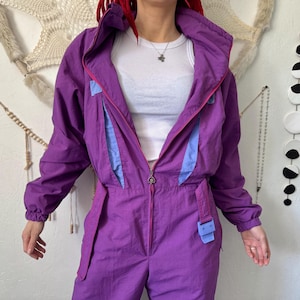 Könnte beinhalten: Ein vintage-violetter Overall mit hohem Kragen, Reißverschluss und hellblauen Akzenten. Das einteilige Kleidungsstück hat eine geschnürte Taille mit Gürtel und Schnalle. Der Overall hat eine leuchtende Farbe und scheint aus Nylon oder Polyester zu sein.