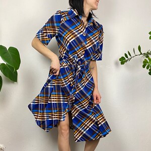 May include: A blue, brown, and white plaid dress with a button-up front and a tie belt. The dress has short sleeves and a knee-length skirt.
