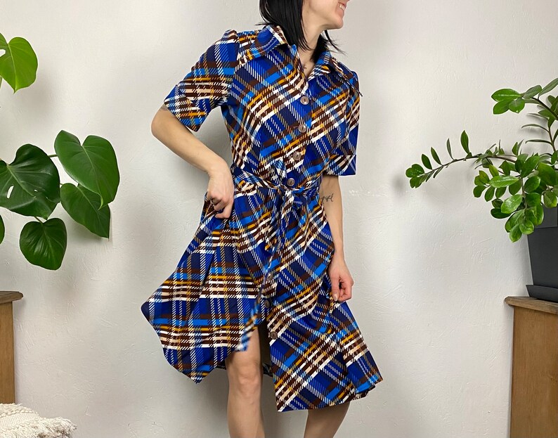 May include: A blue, brown, and white plaid dress with a button-up front and a tie belt. The dress has short sleeves and a knee-length skirt.