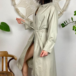 May include: A light beige trench coat with a long sleeve, a button-up front, and a tie belt.