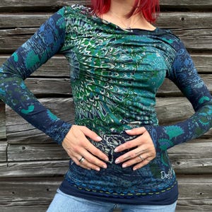 May include: A long-sleeve top featuring a vibrant green and blue peacock feather-inspired pattern with horizontal ruching across the chest. The garment has a round neckline, fitted sleeves, and a dark blue hemline accented with decorative yellow stitching.