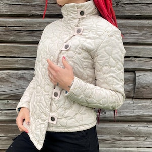 Puede incluir: Chaqueta acolchada color crema con cuello alto y cierre de botones. La chaqueta presenta un patrón único de círculos y óvalos cosidos. Los botones son de color crema a juego. La chaqueta se lleva sobre una prenda negra.