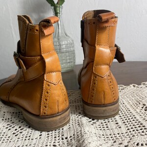 K&ouml;nnte beinhalten: Ein Paar hellbraune Leder-Ankle Boots mit dekorativen Perforationen und Schnallenverschl&uuml;ssen. Die Stiefel haben einen niedrigen Absatz und ein klassisches Design, das f&uuml;r verschiedene Outfits geeignet ist. Die Stiefel werden auf einer wei&szlig;en Spitzentischdecke pr&auml;sentiert.