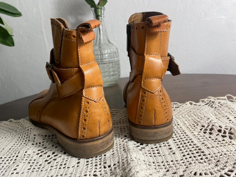 K&ouml;nnte beinhalten: Ein Paar hellbraune Leder-Ankle Boots mit dekorativen Perforationen und Schnallenverschl&uuml;ssen. Die Stiefel haben einen niedrigen Absatz und ein klassisches Design, das f&uuml;r verschiedene Outfits geeignet ist. Die Stiefel werden auf einer wei&szlig;en Spitzentischdecke pr&auml;sentiert.