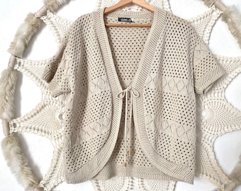 May include: A beige crochet cardigan with a tie front and short sleeves. The cardigan has a delicate openwork pattern.