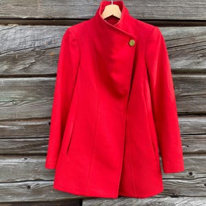 May include: A vibrant red coat with a high collar and a single gold button closure. The coat is hanging on a wooden hanger against a weathered wood background. The coat's design features a wrap-style front.