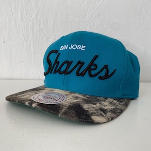 Può includere: Un cappellino da baseball color verde acqua con la scritta SAN JOSE in bianco e Sharks ricamato in corsivo nero. La visiera piatta presenta un motivo tie-dye bianco e nero e un adesivo circolare con la scritta Mitchell & Ness Adjustable Fit.