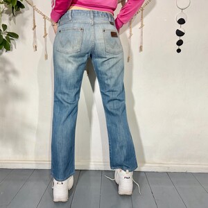 May include: Light blue denim jeans with a faded wash. The jeans feature a classic five-pocket design, a brown leather patch, and the Wrangler logo. The jeans have a slight bootcut and are worn with white sneakers.