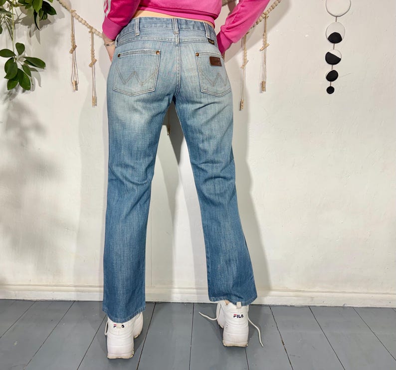 May include: Light blue denim jeans with a faded wash. The jeans feature a classic five-pocket design, a brown leather patch, and the Wrangler logo. The jeans have a slight bootcut and are worn with white sneakers.