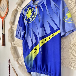 Puede incluir: Maillot de ciclismo azul y celeste con un logotipo de escorpión amarillo y la palabra "Castelli". El maillot tiene una cremallera parcial y la palabra "SYSTEM" en amarillo. El maillot es de manga corta y cuelga de una percha de madera.