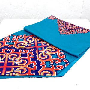 May include: Two teal-colored decorative fabric pieces. One piece is folded, revealing a patterned design in orange, red, and blue. The other piece has a similar design in the corner. The fabric is likely used for home decor.