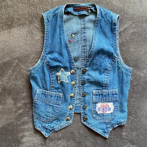 Può includere: Un gilet in denim blu con scollo a V, chiusura frontale con bottoni metallici e due tasche inferiori. Il gilet presenta una toppa sfrangiata a forma di stella sul lato destro e una toppa rettangolare con testo rosso e blu sulla tasca sinistra.