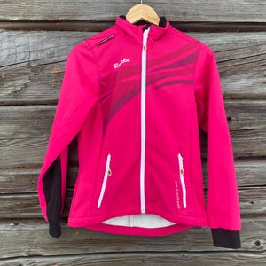 Könnte beinhalten: Leuchtend pinke Jacke mit weißem Reißverschluss und Details. Schwarze Ärmel und Markenname "Rukka" sichtbar. Auf einem Holzbügel vor Holzgrund. Ideal für Outdoor-Aktivitäten.