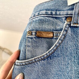 May include: Close-up of light blue denim jeans. The jeans feature a small pocket with a brown leather label. The label has the brand name. The jeans have a classic five-pocket design with copper-colored rivets and stitching.