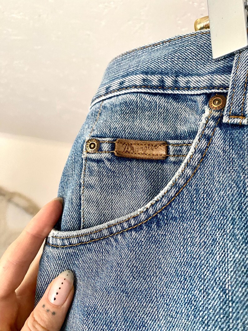 May include: Close-up of light blue denim jeans. The jeans feature a small pocket with a brown leather label. The label has the brand name. The jeans have a classic five-pocket design with copper-colored rivets and stitching.