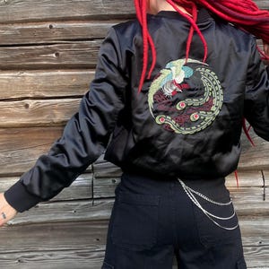 May include: Black satin bomber jacket with an embroidered phoenix design on the back. The jacket has a ribbed collar and cuffs. The model is wearing black trousers and a silver chain.