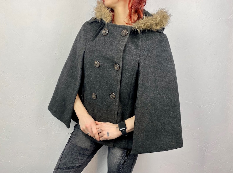 May include: A charcoal gray cape coat with a faux fur-trimmed hood. The double-breasted coat features large, round buttons and a classic design. The cape has a relaxed fit and is made of a soft, wool-like material.