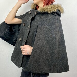 May include: A charcoal gray cape with a faux fur-trimmed hood. The cape features a button-up front and a black lining. The garment is made of a wool-like material, offering a classic and warm outerwear option.