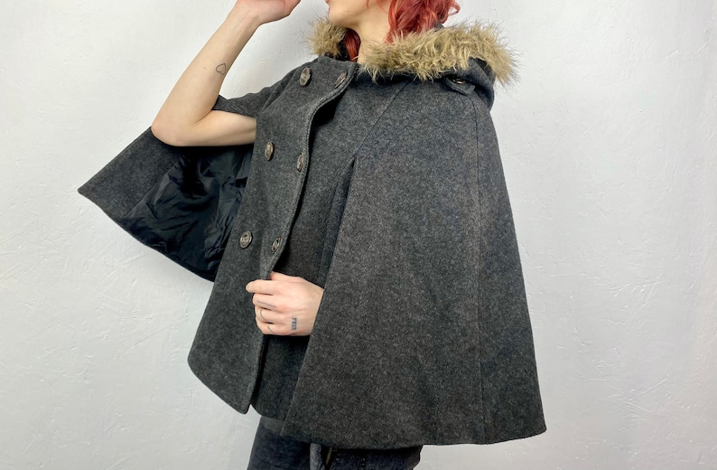 May include: A charcoal gray cape with a faux fur-trimmed hood. The cape features a button-up front and a black lining. The garment is made of a wool-like material, offering a classic and warm outerwear option.
