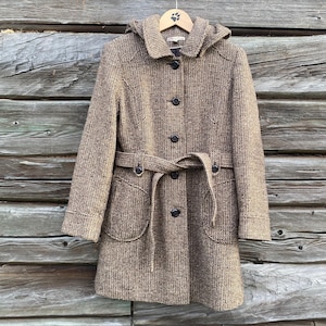 Può includere: Un cappotto marrone in tweed con cappuccio, chiusura con bottoni e cintura. Il cappotto ha due tasche anteriori ed è appeso a una gruccia di legno su uno sfondo di legno.