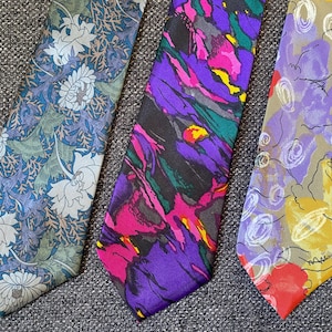 Könnte beinhalten: Drei Seidenkrawatten mit verschiedenen abstrakten Mustern. Eine Krawatte hat ein Blumenmuster in Blautönen und Grün, eine andere hat ein wirbelndes Muster in Violett-, Rosa- und Gelbtönen, und die dritte hat ein abstrakteres Muster in Violett-, Gelb- und Rottönen.