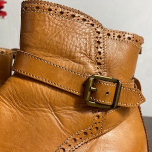 K&ouml;nnte beinhalten: Nahaufnahme eines hellbraunen Lederstiefels mit dekorativem Riemen und Schnalle. Der Stiefel hat ein detailliertes, perforiertes Design entlang der Oberkante und um den Kn&ouml;chel. Das Leder wirkt abgenutzt und strukturiert, mit sichtbaren N&auml;hten.