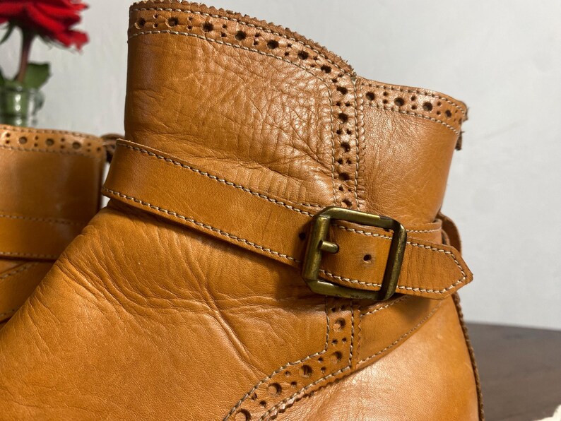 K&ouml;nnte beinhalten: Nahaufnahme eines hellbraunen Lederstiefels mit dekorativem Riemen und Schnalle. Der Stiefel hat ein detailliertes, perforiertes Design entlang der Oberkante und um den Kn&ouml;chel. Das Leder wirkt abgenutzt und strukturiert, mit sichtbaren N&auml;hten.