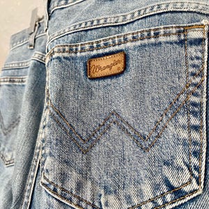May include: Light blue denim jeans with a classic five-pocket design. The back pocket features a signature Wrangler stitching pattern and a brown leather patch with the brand name. The jeans are hanging on a wooden hanger.