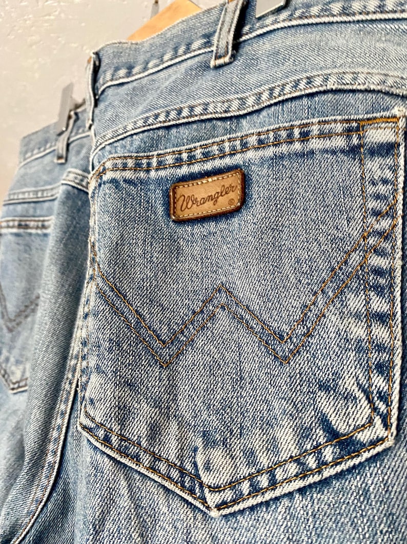 May include: Light blue denim jeans with a classic five-pocket design. The back pocket features a signature Wrangler stitching pattern and a brown leather patch with the brand name. The jeans are hanging on a wooden hanger.